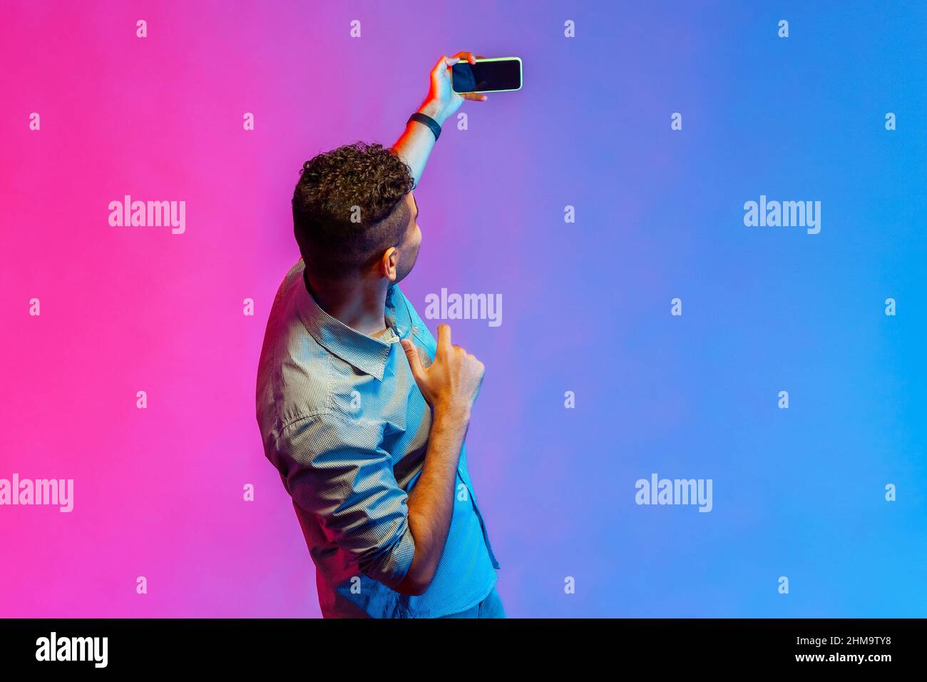 Back side of man in shirt taking selfie or broadcasting livestream, holding smart phone with blank screen, online conversation. Indoor studio shot isolated on colorful neon light background. Stock Photo