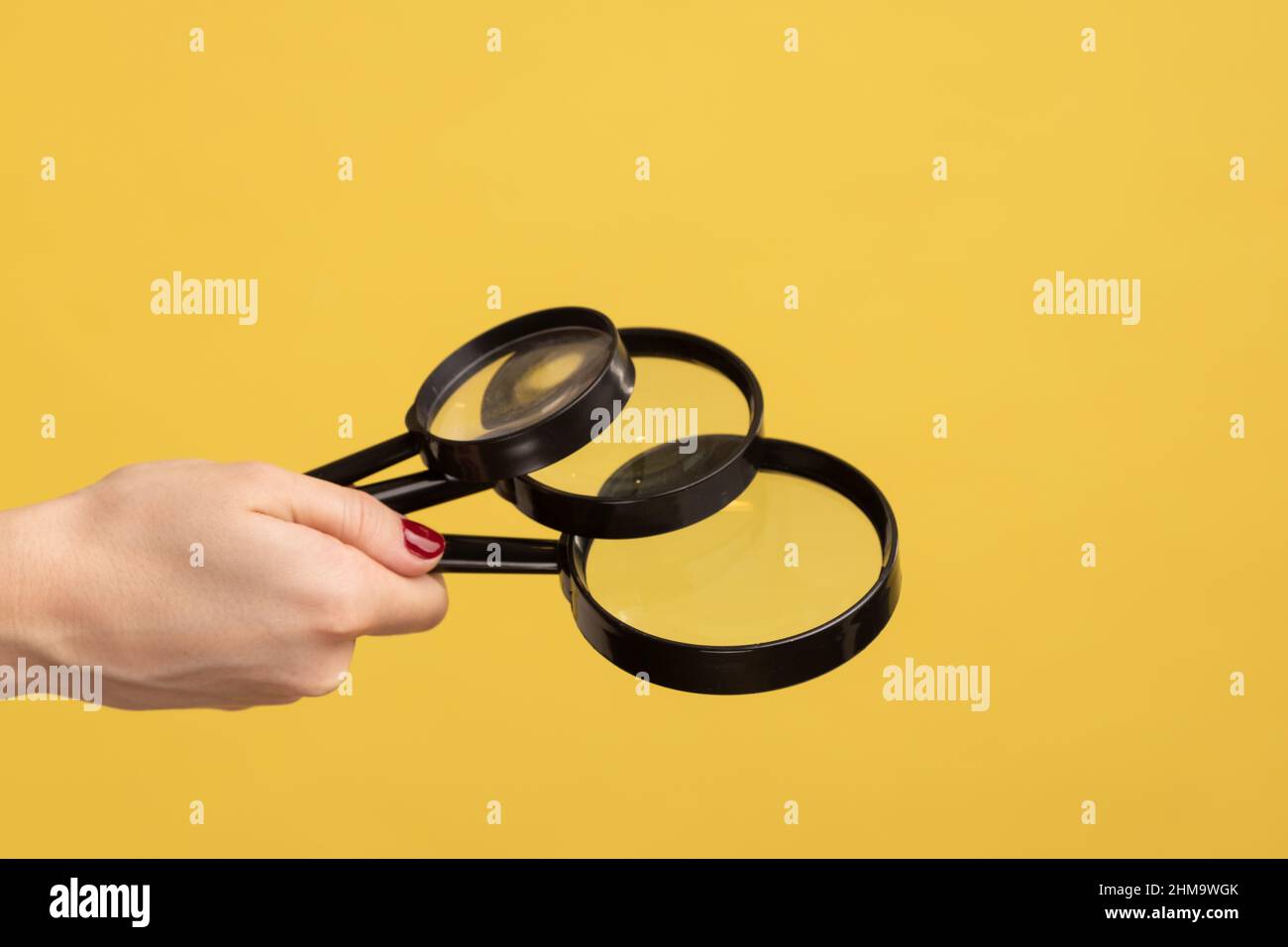 Profile side view closeup of woman hand holding several magnify glasses, loupe. Indoor studio shot isolated on yellow background. Stock Photo
