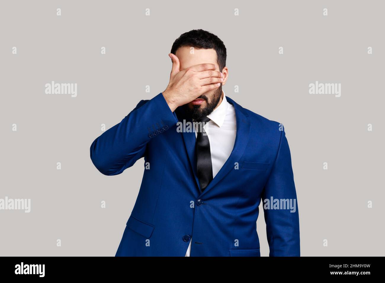 Bearded man closing eyes with hand, dont want to see that, ignoring problems, hiding from stressful situations, wearing official style suit. Indoor studio shot isolated on gray background. Stock Photo