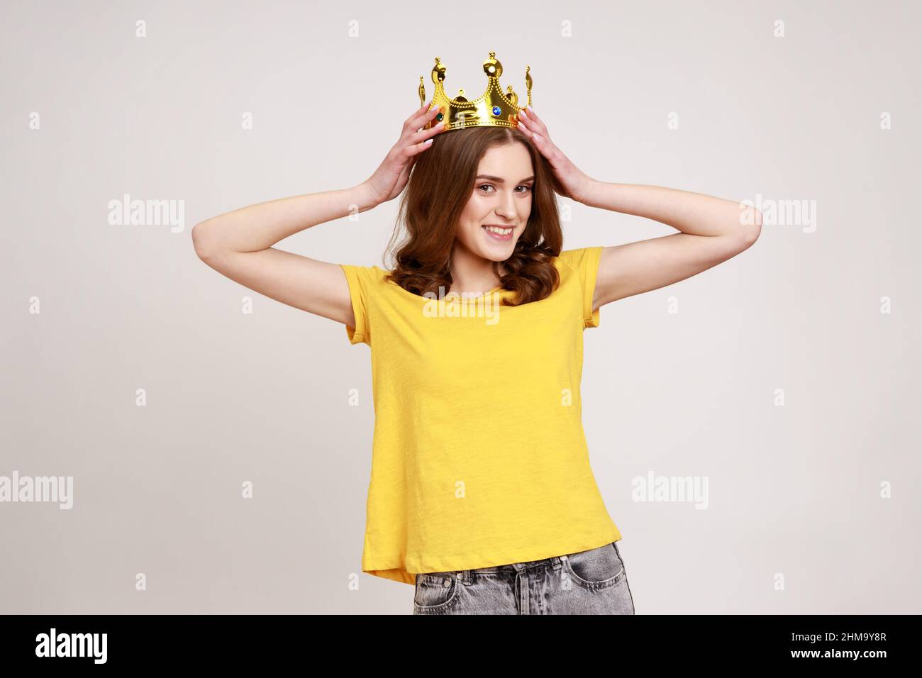 I am queen. Portrait of happy curly-haired teenager girl putting crown on head and smiling, self confidence, self-motivation and dreams to be best. Indoor studio shot isolated on gray background. Stock Photo