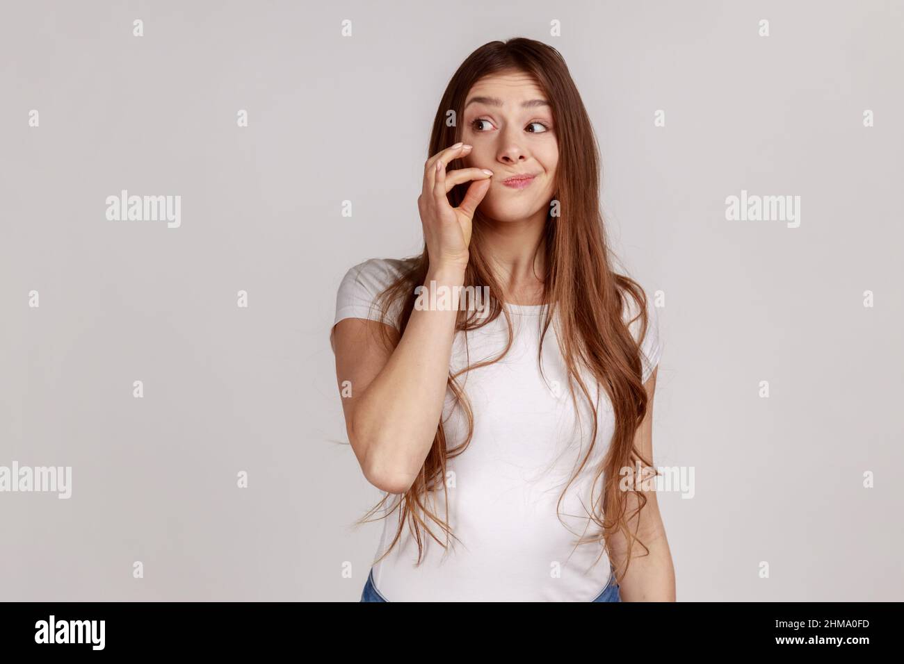 Attractive woman with long dark hair and mystery look making zip gesture to close mouth, keeping secret, zipping lips, wearing white T-shirt. Indoor studio shot isolated on gray background. Stock Photo