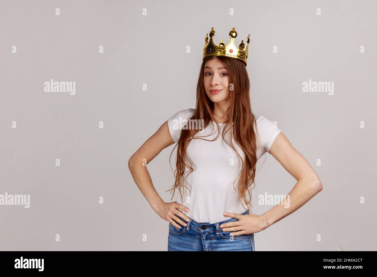 Portrait of young beautiful woman with crown on head keeping hands on hips, self-motivation and dreams to be best, wearing white T-shirt. Indoor studio shot isolated on gray background. Stock Photo
