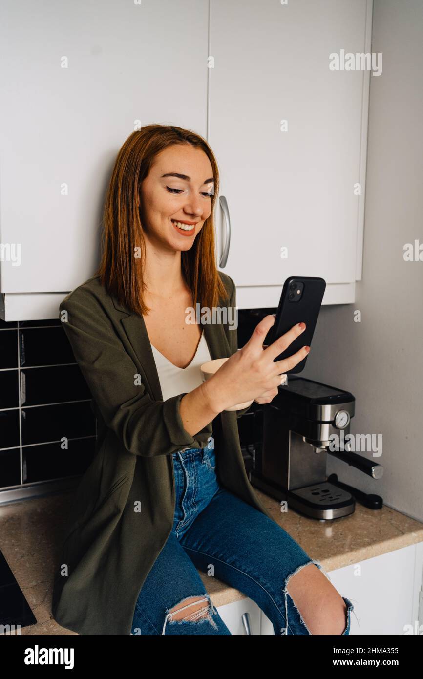 Focused female with red hair in ripped jeans sitting on kitchen counter with cup of coffee and browsing smartphone Stock Photo