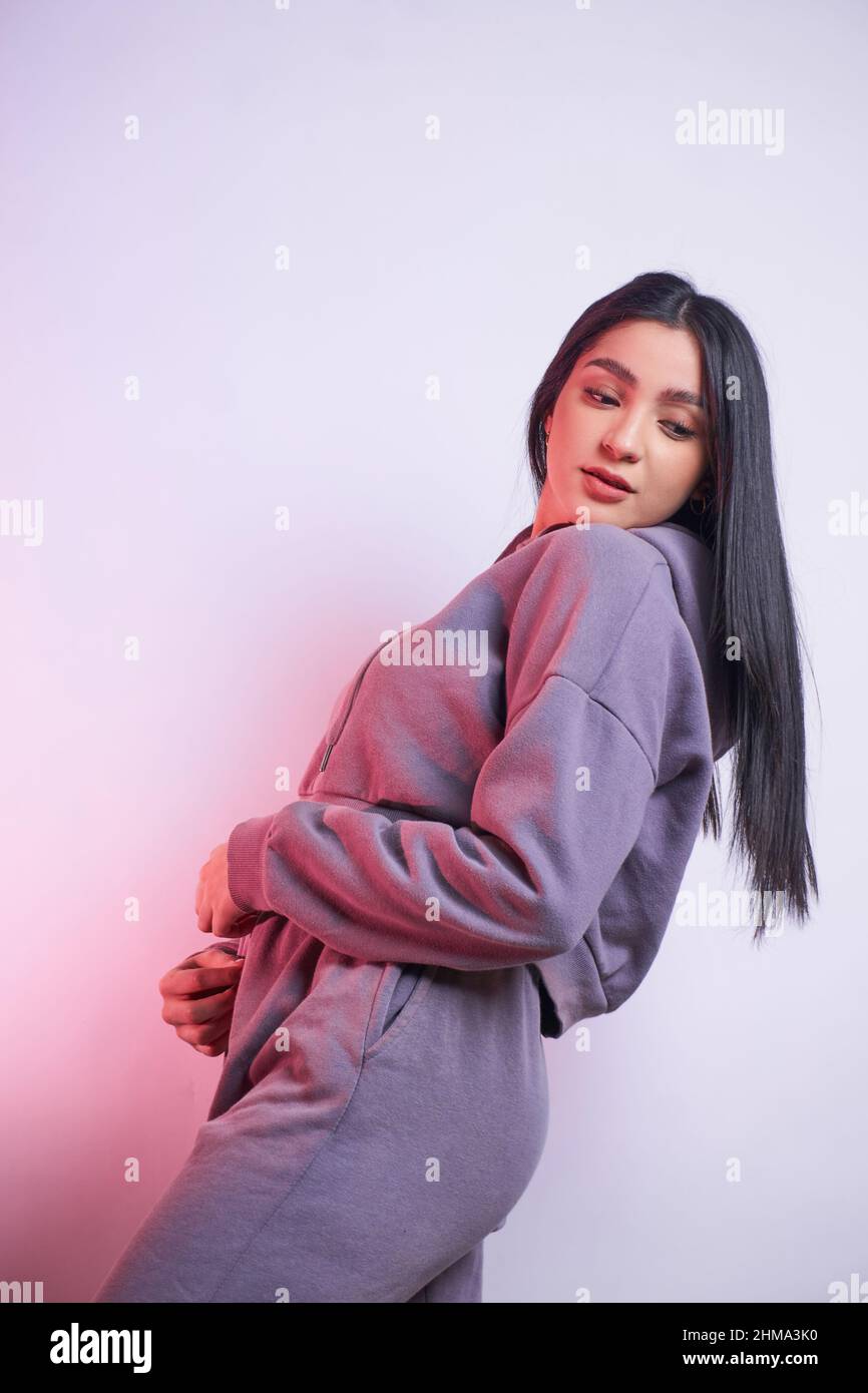 Low angle young female model with black hair in joggers and hoodie set looking down over shoulder while standing on light background in studio Stock Photo