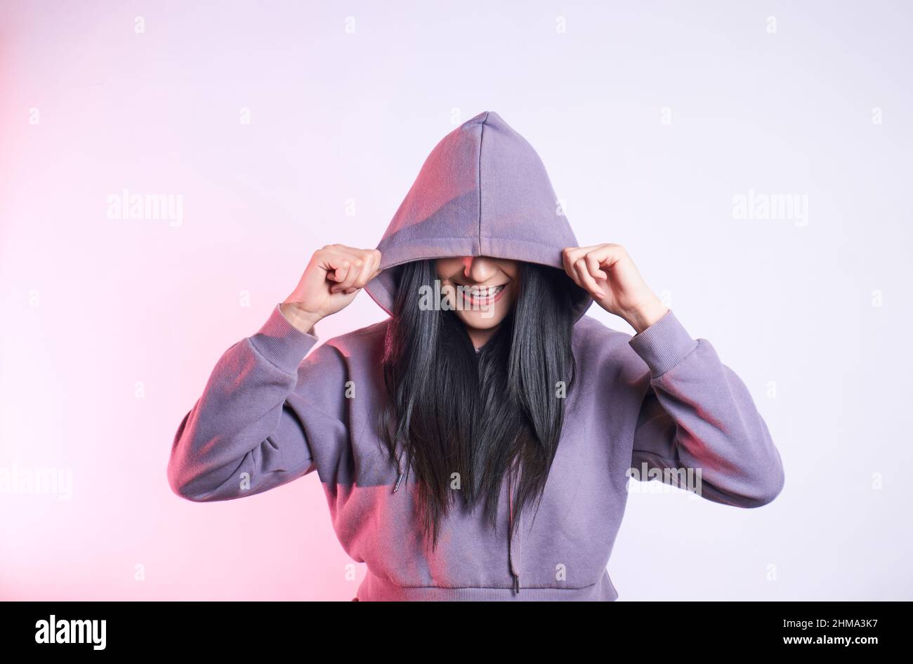 Cheerful unrecognizable female with black hair covering eyes with casual hoodie while standing on purple background in modern light studio Stock Photo