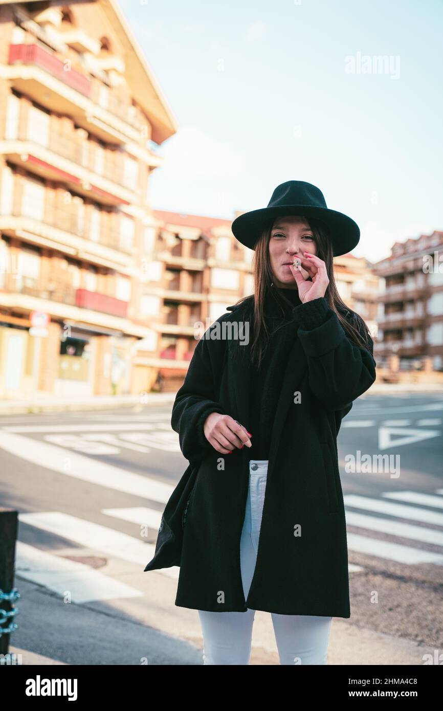 Content young female in black hat looking at camera while smoking cigarette near asphalt road on street with residential buildings Stock Photo