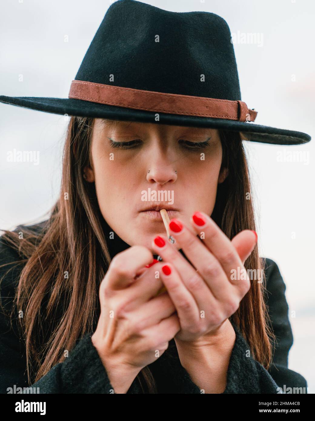 Attractive female smoker with long brown hair in stylish black headwear lighting cigarette while smoking on street against cloudless sky Stock Photo