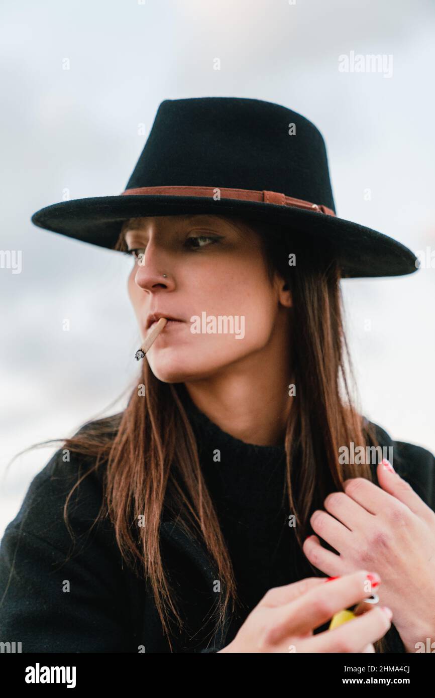 Attractive female smoker with long brown hair in stylish black headwear smoking cigarette on street against cloudless sky Stock Photo