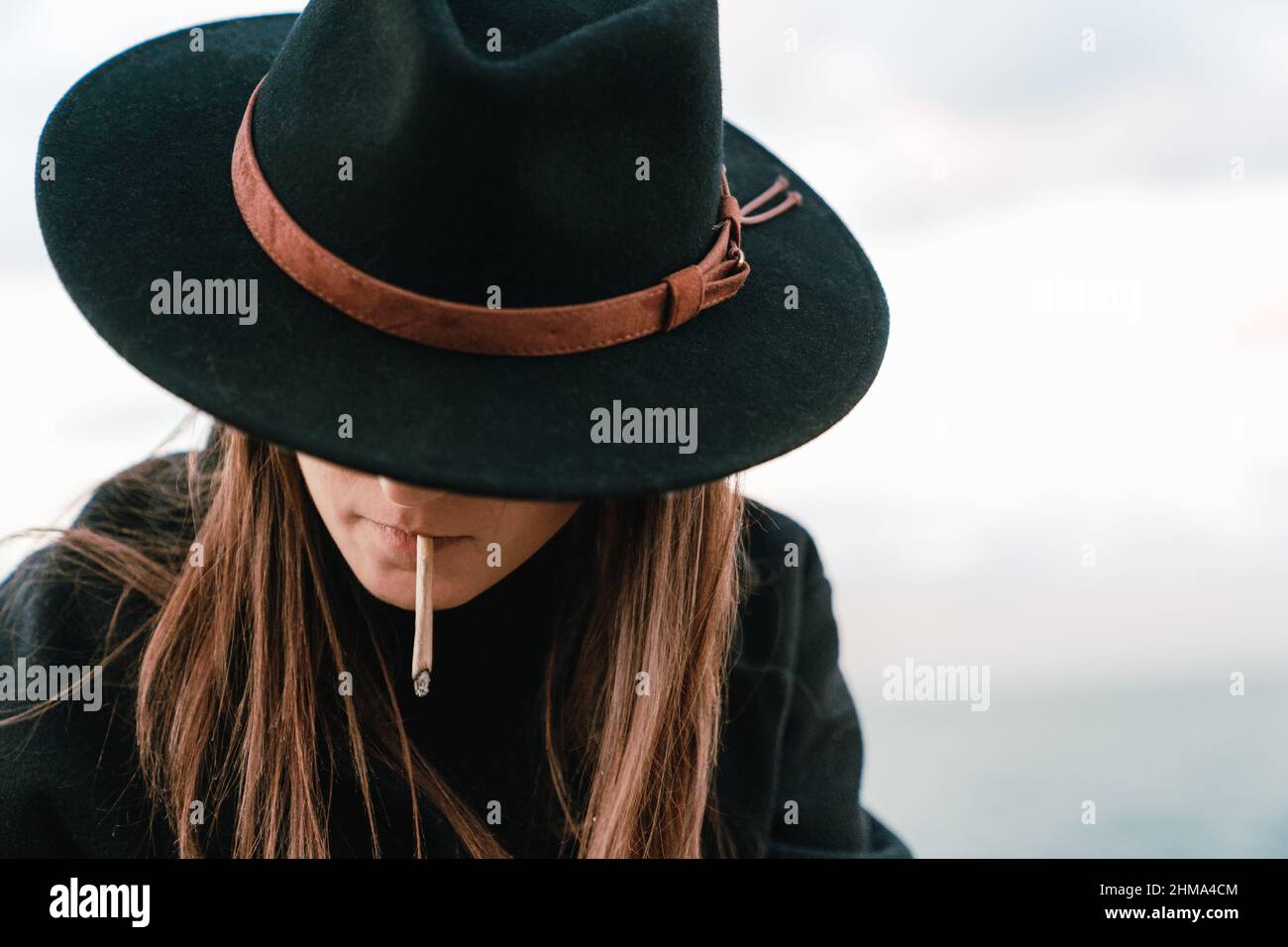 Unrecognizable female smoker with long brown hair in stylish black headwear smoking cigarette on street against cloudless sky Stock Photo