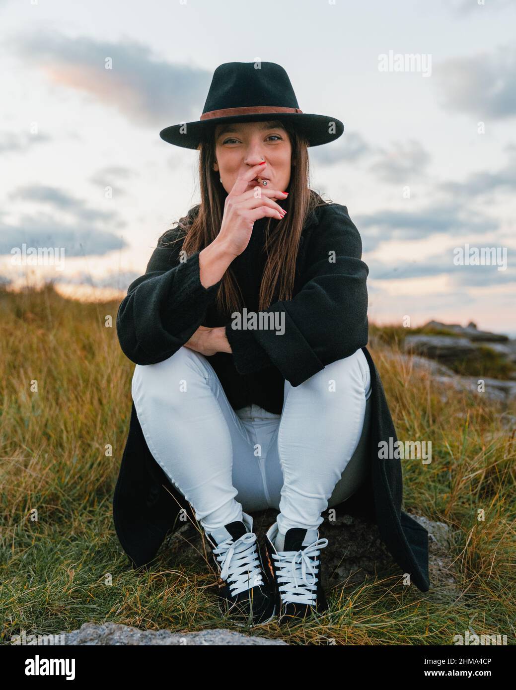 Attractive smiling female smoker with long brown hair in stylish black headwear smoking cigarette on street against cloudless sky on meadow Stock Photo