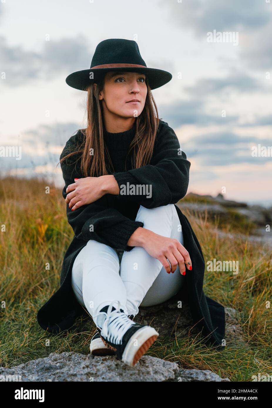 Attractive female smoker with long brown hair in stylish black headwear smoking cigarette on street against cloudless sky on meadow Stock Photo