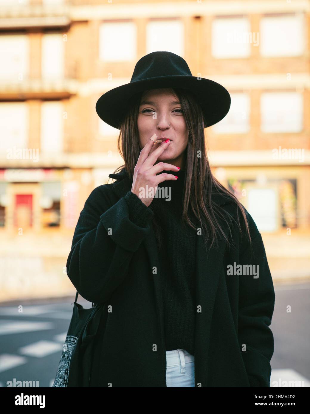 Content young female in black hat looking at camera while smoking cigarette near asphalt road on street with residential buildings Stock Photo