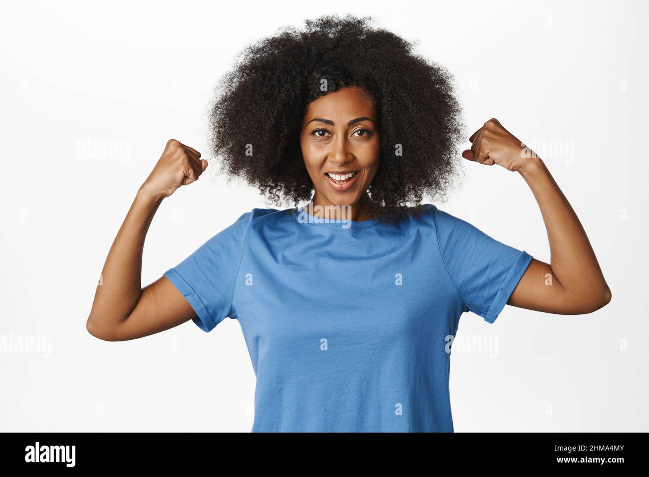 Women power. Enthusiastic african american woman flexing biceps, showing strong muscles, feeling strength and looking confident, standing in blue t Stock Photo