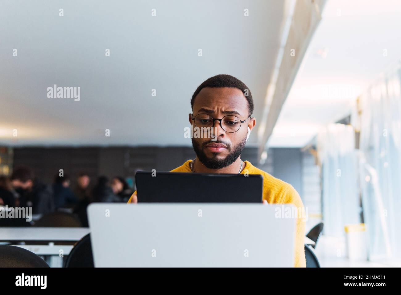 Thoughtful black male sitting at table with laptop and listening for music in earphones from tablet in coworking space Stock Photo