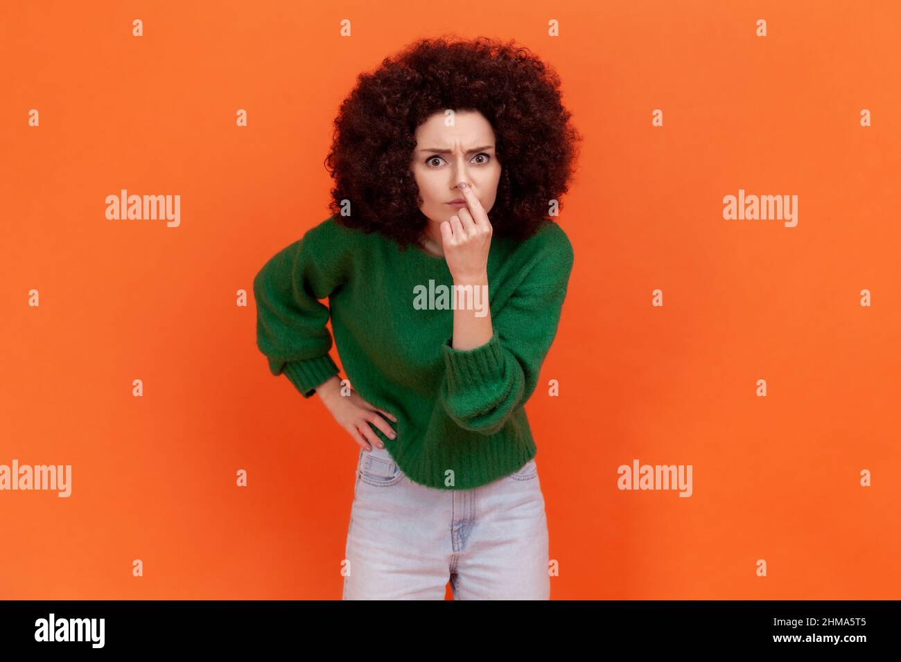 Woman with Afro hairstyle looking with incredulous suspicious gaze and touching nose, gesturing you are liar, suspecting falsehood. Indoor studio shot isolated on orange background. Stock Photo