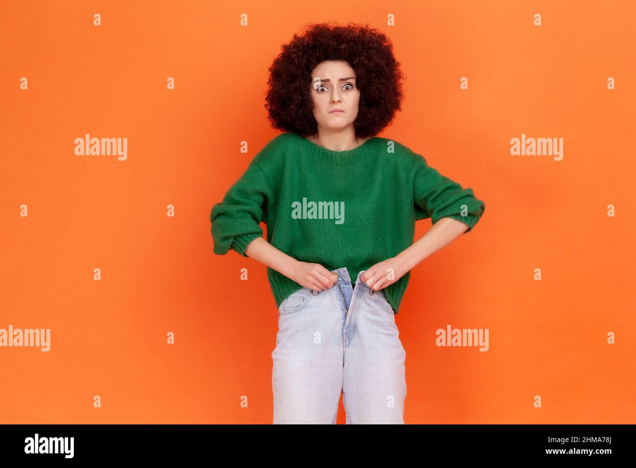 Portrait of upset sad young adult woman with Afro hairstyle wearing green casual style sweater unable to button jeans due to weight gain. Indoor studio shot isolated on orange background. Stock Photo