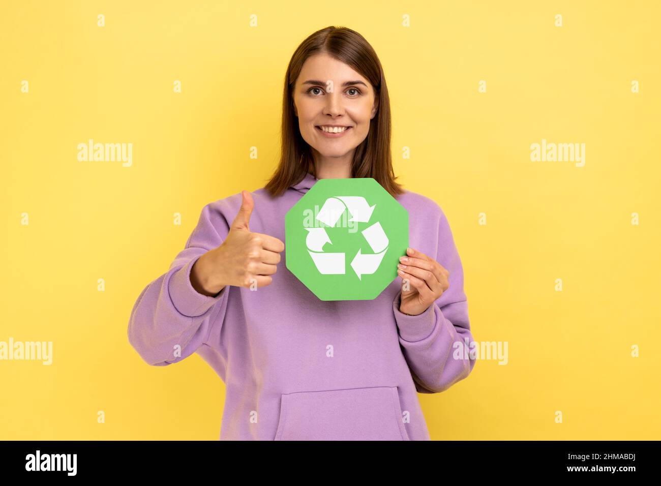 Positive woman with dark hair holding in hands green recycling sing, ecology concept, looking at camera and showing thumb up, wearing purple hoodie. Indoor studio shot isolated on yellow background. Stock Photo