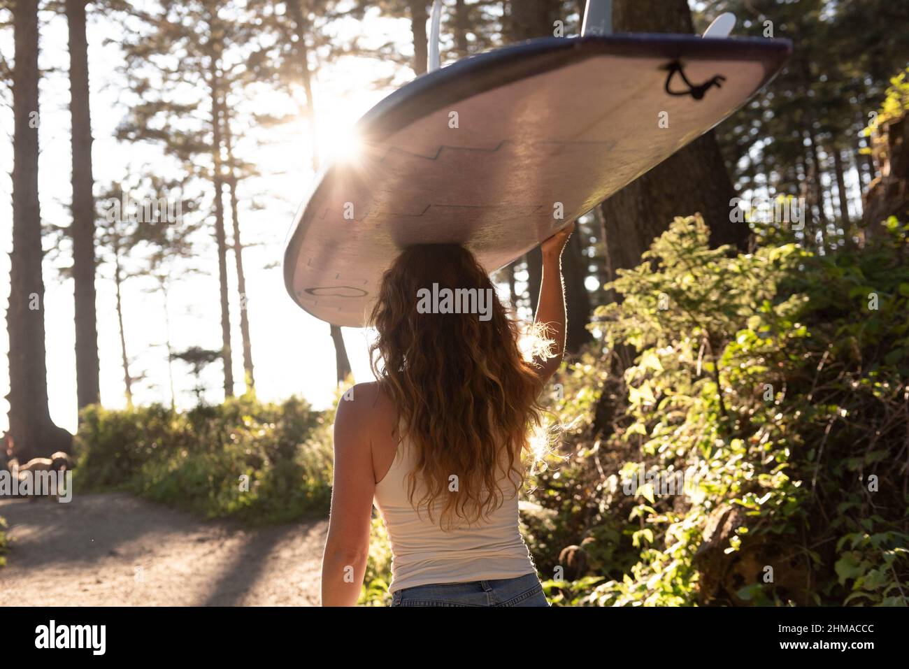 Woman carrying surfboard on her head down trail to the ocean Stock