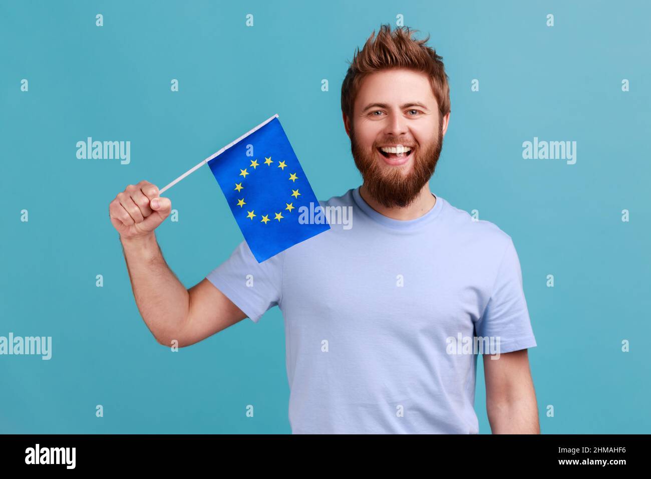 Portrait of excited bearded man smiling broadly and holding flag of European Union, symbol of Europe, EU association and community. Indoor studio shot isolated on blue background. Stock Photo