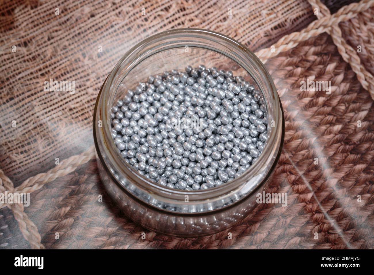 Silver sugar dragee in a plastic bowl. It is on a table covered with a patterned tablecloth. Close-up. Stock Photo