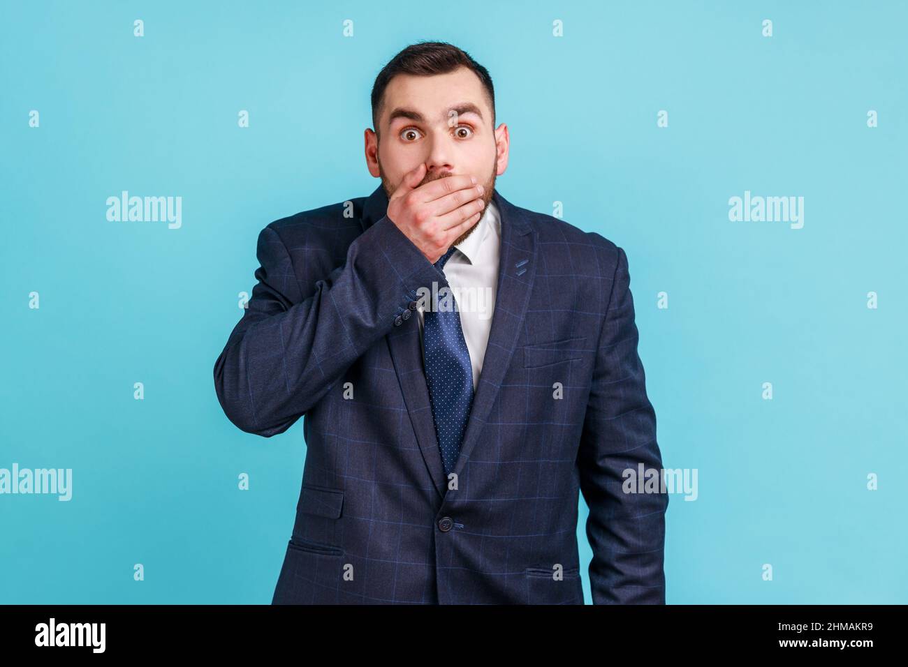 I won't tell. Portrait of amazed businessman wearing official style suit covering mouth with hand, keeping secret, terrified with shocking news. Indoor studio shot isolated on blue background. Stock Photo