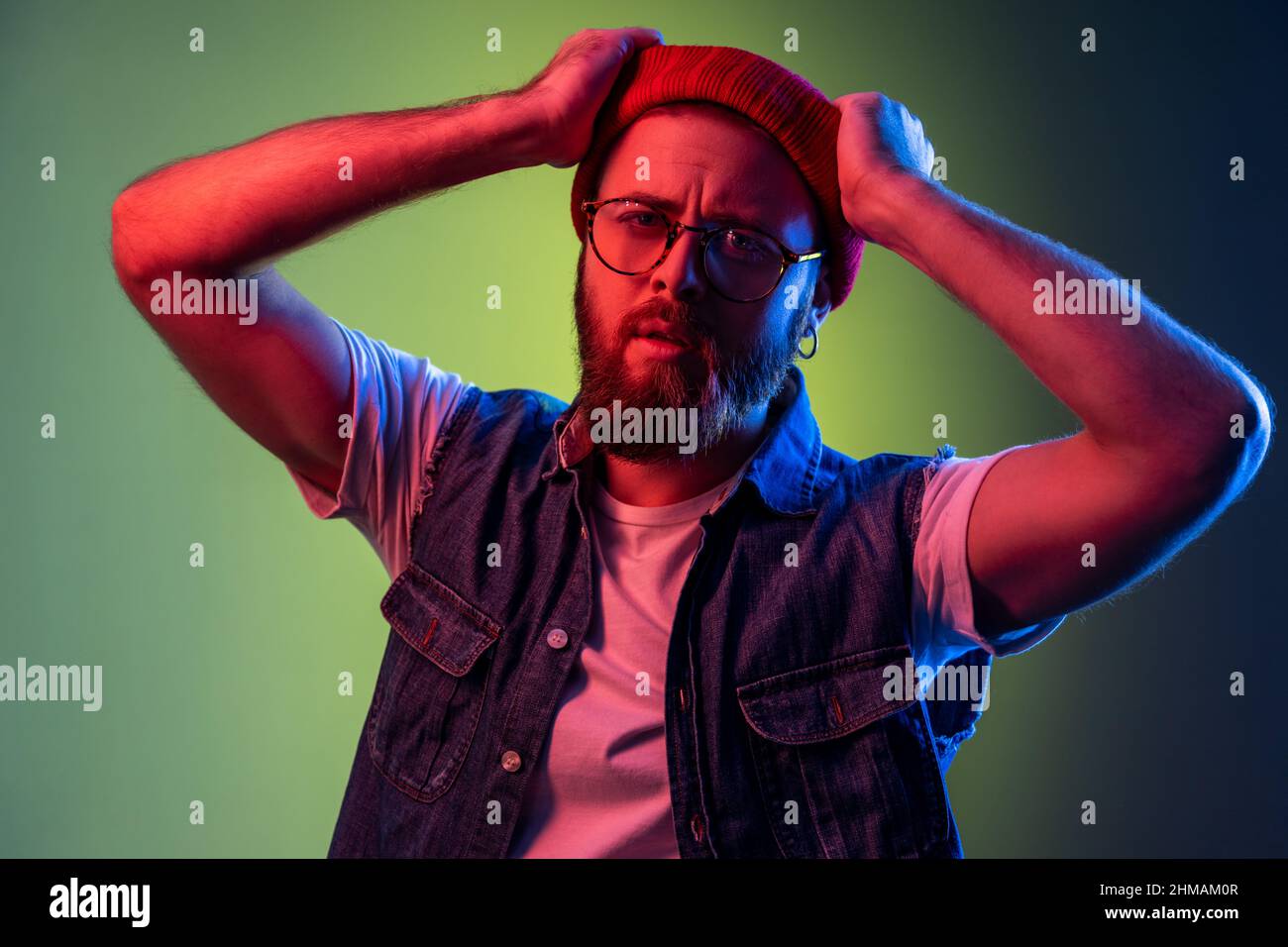 Young adult bearded hipster man in glasses, looking at camera, raised arms, expressing pensive dreamy emotions, wearing red beanie hat. Indoor studio shot isolated on colorful neon light background. Stock Photo