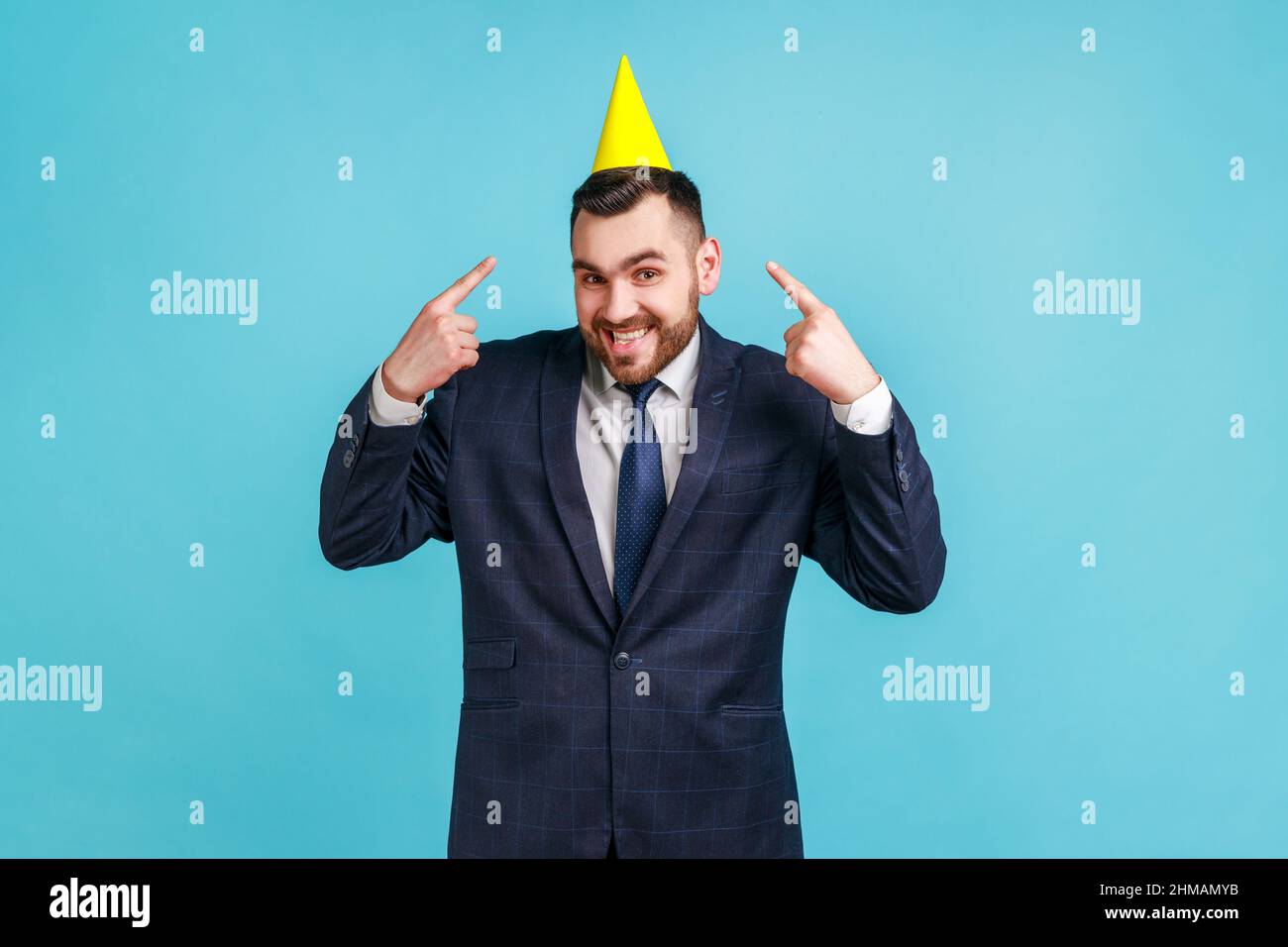 Happy birthday to me. Portrait of cheerful handsome man wearing dark suit man pointing at funny party hat and looking at camera with toothy smile. Indoor studio shot isolated on blue background. Stock Photo