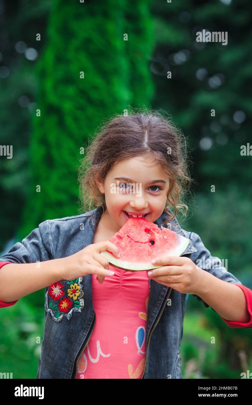 Little happy girl eating a juicy watermelon in the garden. Children eat fruit outdoors. Healthy food for children. Stock Photo