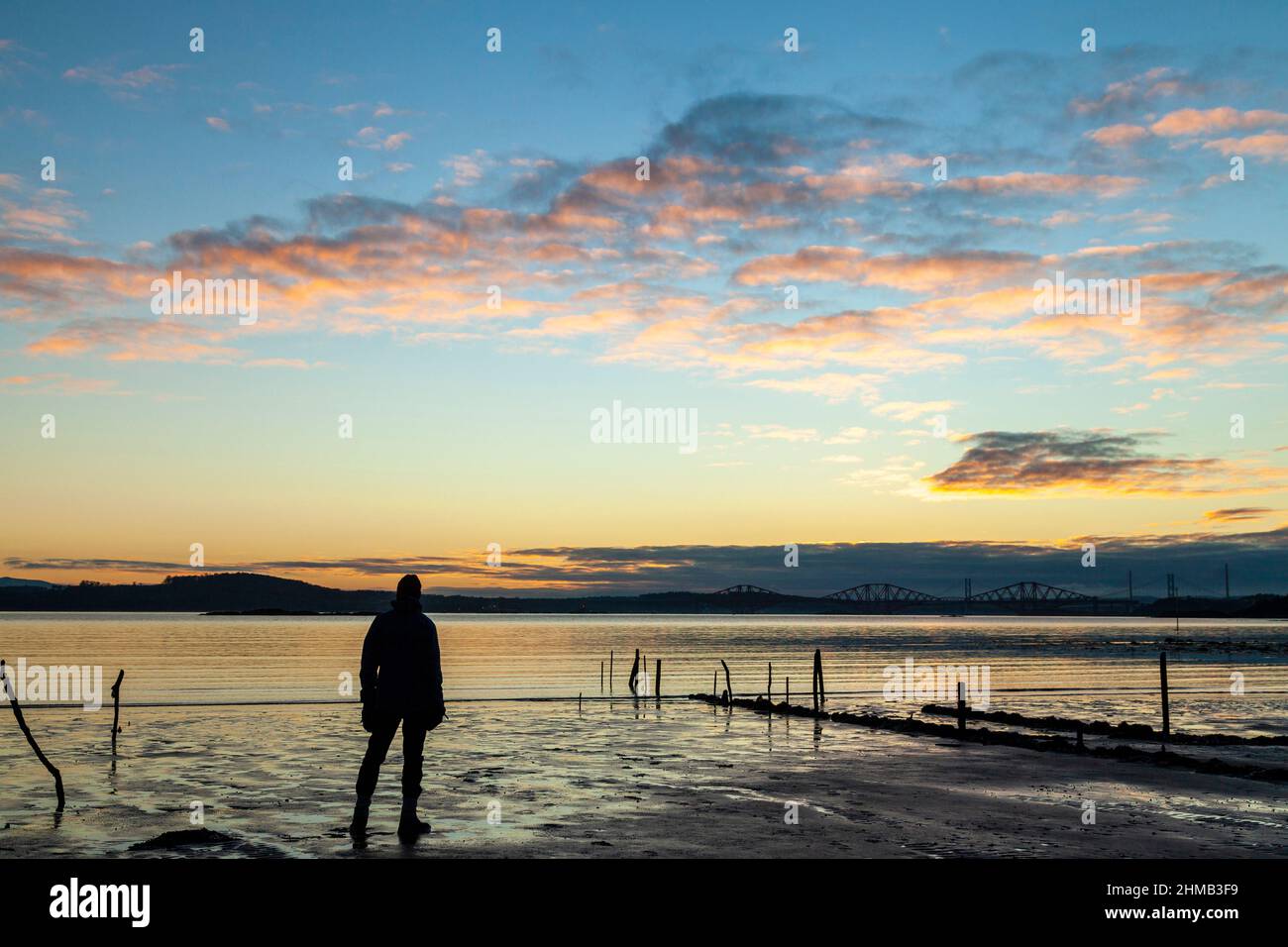 A person looking at the sunrise over the Firth of Forth in Dalgety Bay with the Forth Bridge in the distance. Stock Photo