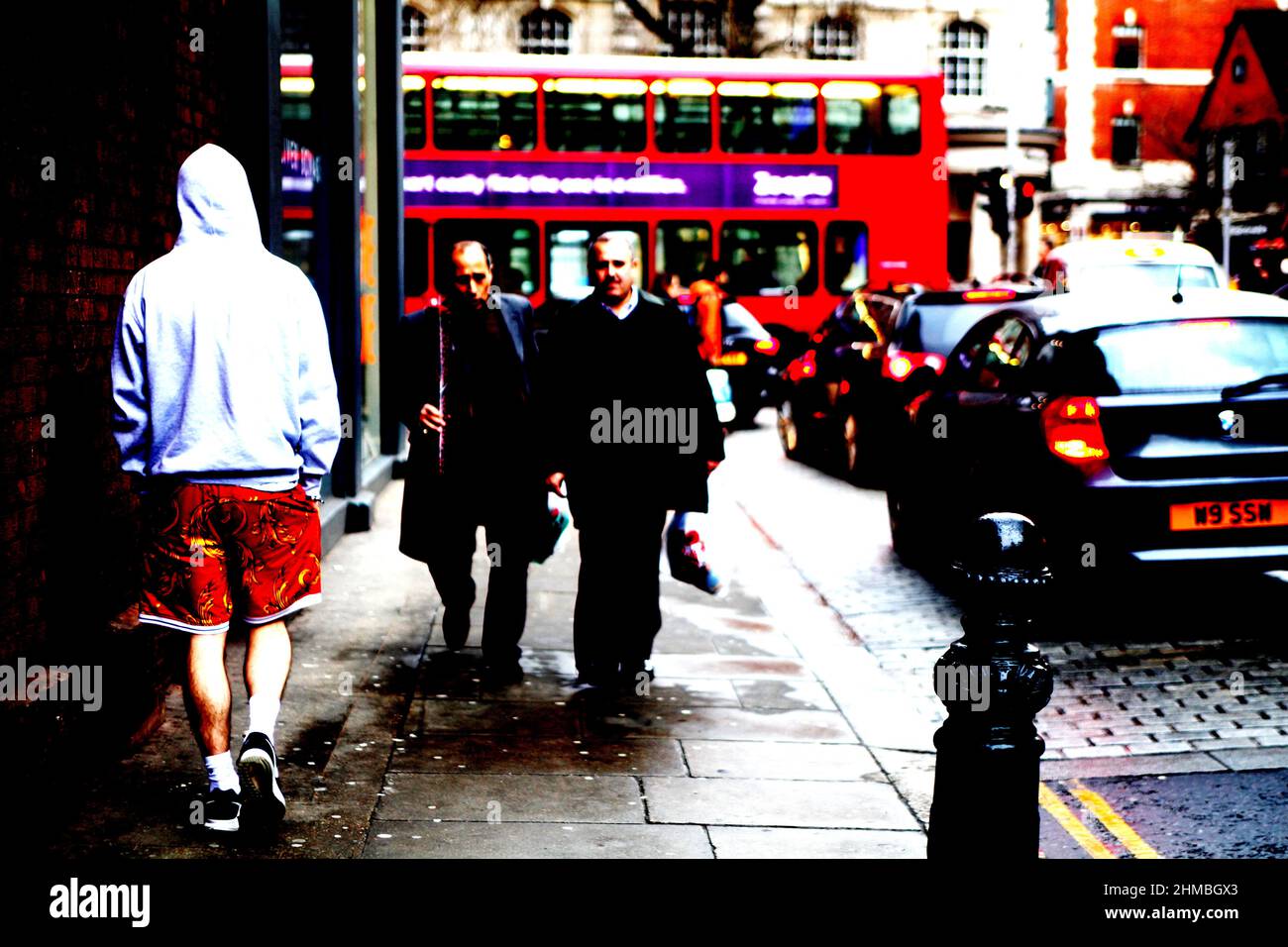 Young man in big red shorts walking on the street in London with two middle age gentlemen in casual suits walk his side on a grey St Valentine day.. Stock Photo