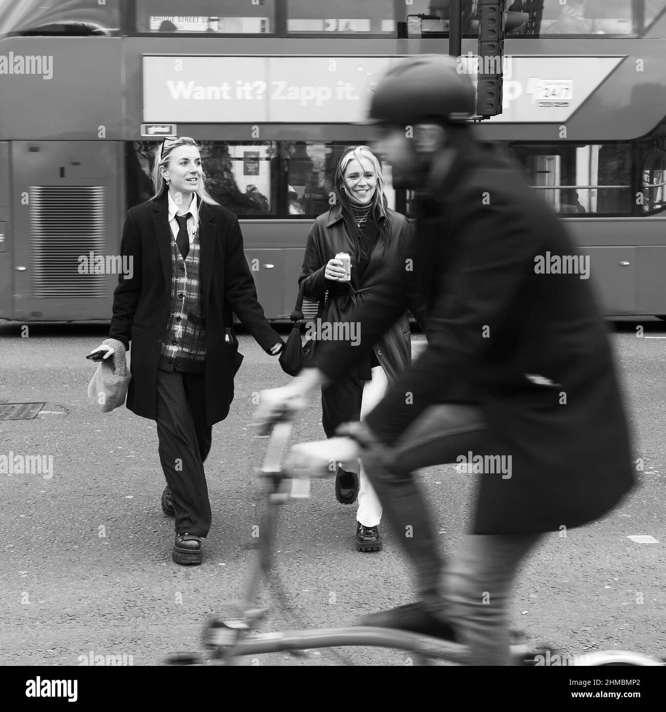 Two trendy women one wearing a waistcoat smiling and smirking walk across Bridge Street as a cyclist, London. Stock Photo