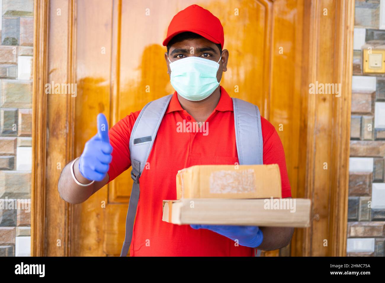 Focus on face, delivery boy with medical face mask and cardboard boxes in hands showing thumbs up gesture bylooking at camera - concept of online Stock Photo