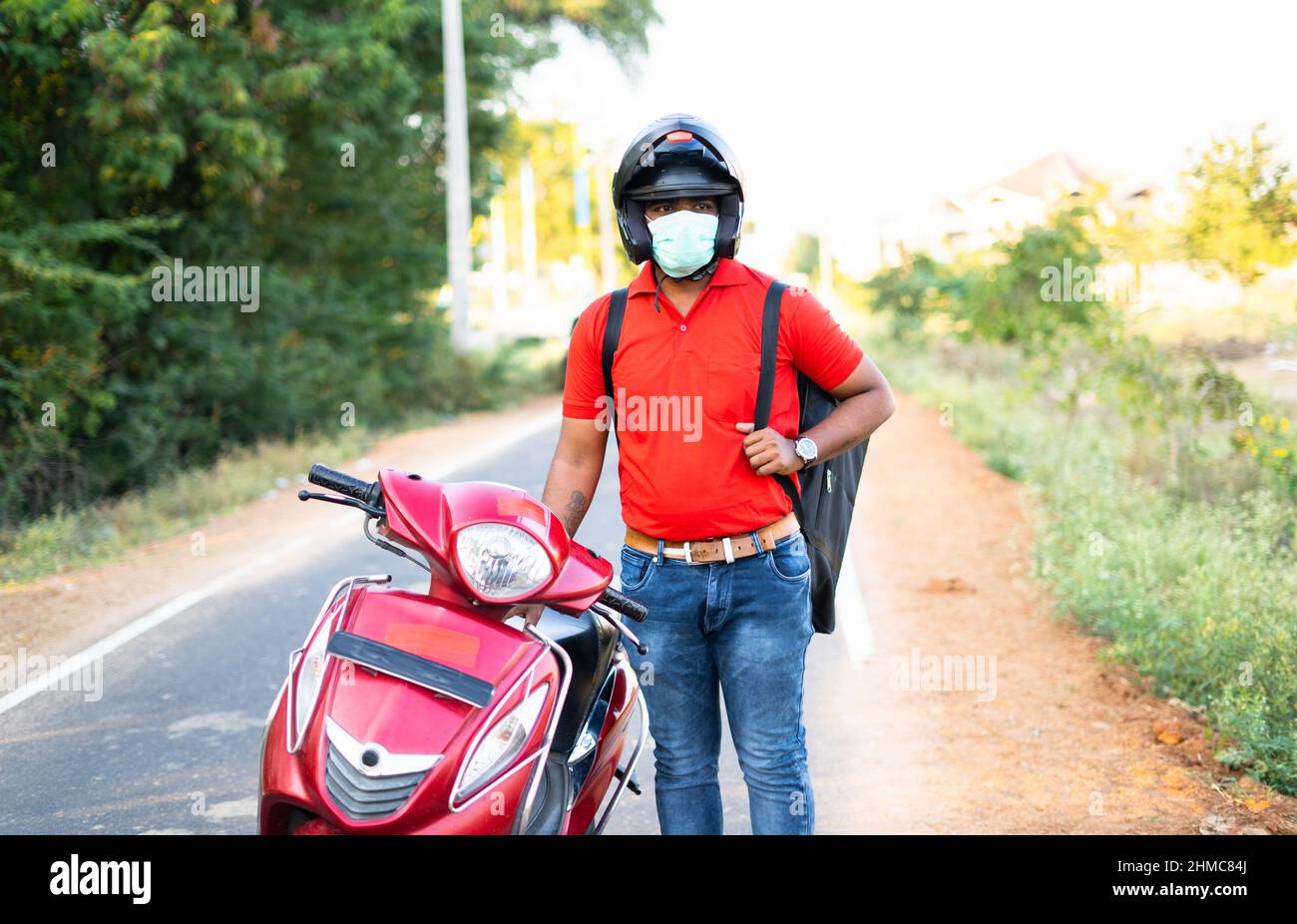 Delivery boy medical face mask coming by standing next to scooter - concept of courier service during coronavirus or covid-19 pandemic with safety Stock Photo