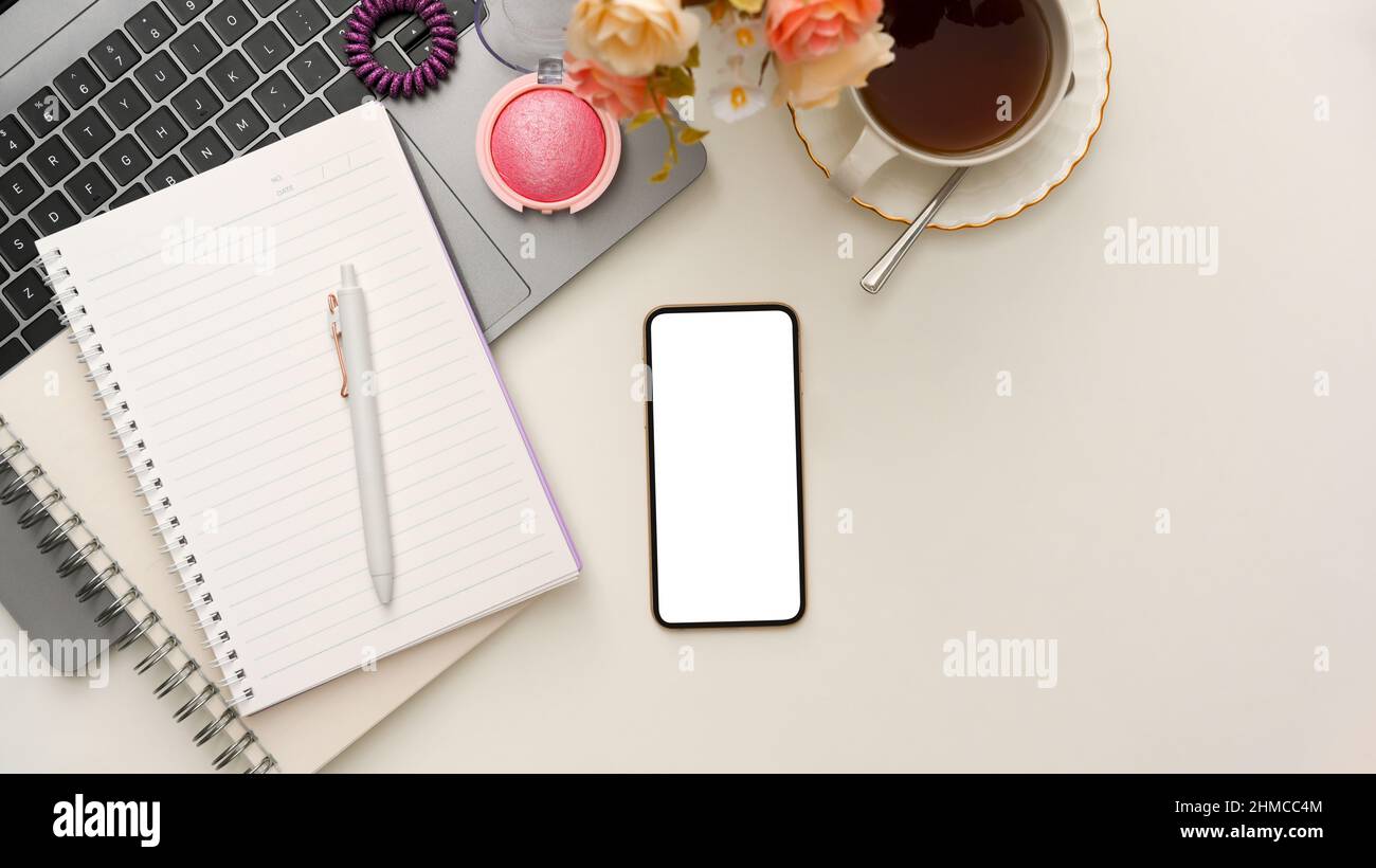 Overhead, Minimal feminine workspace or office desk with spiral notebook, teacup, laptop, makeup, smartphone white screen mockup and copy space on whi Stock Photo