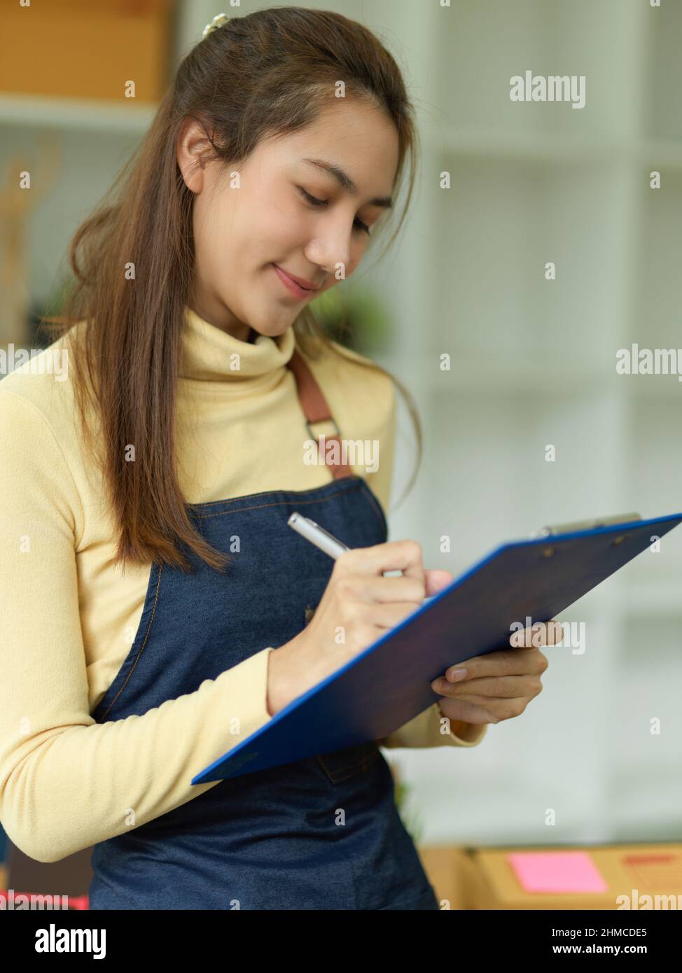 Portrait of a beautiful asian female waitress taking a food and drink orders on clipboard paper. Friendly server wears apron and takes orders in resta Stock Photo