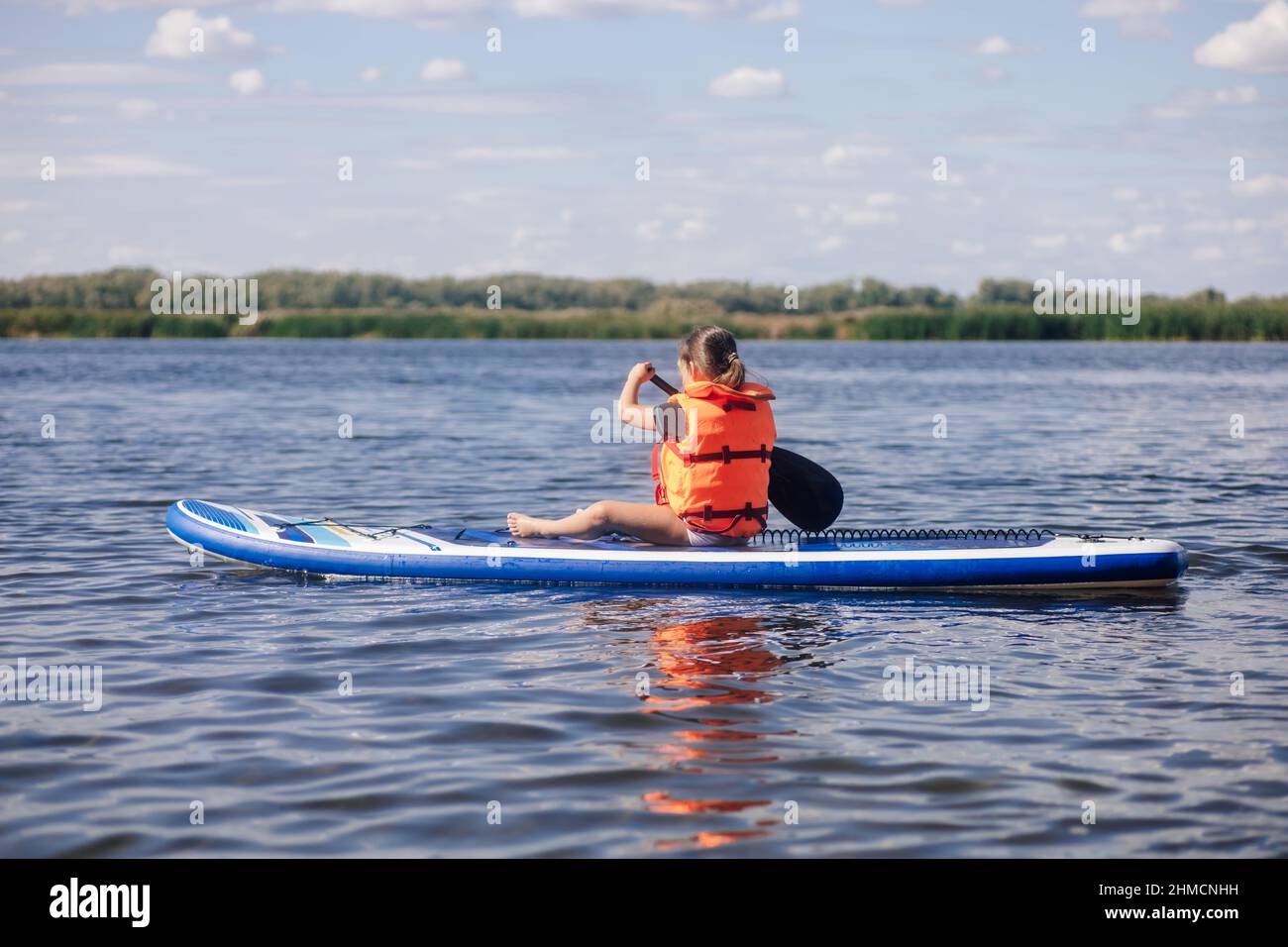 Blonde small girl sup boarding on lake in daytime rowing with oar with reeds and trees in background wearing vest life jacket. Active holidays Stock Photo