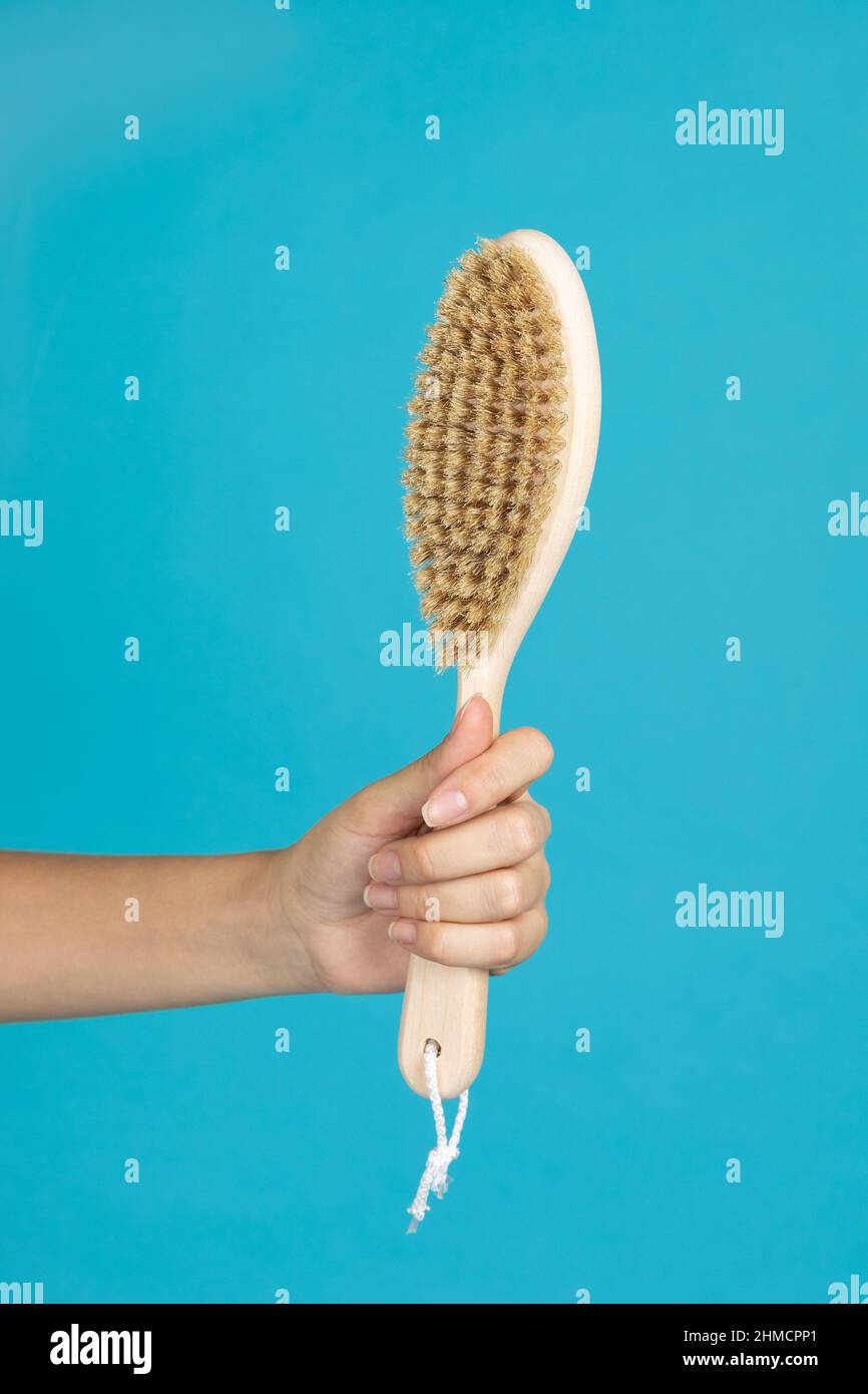 Closeup of hand holding wooden big brush for dry skin massage on blue background. Anti-cellulite procedures aiming for getting rid of cellulite. Self Stock Photo