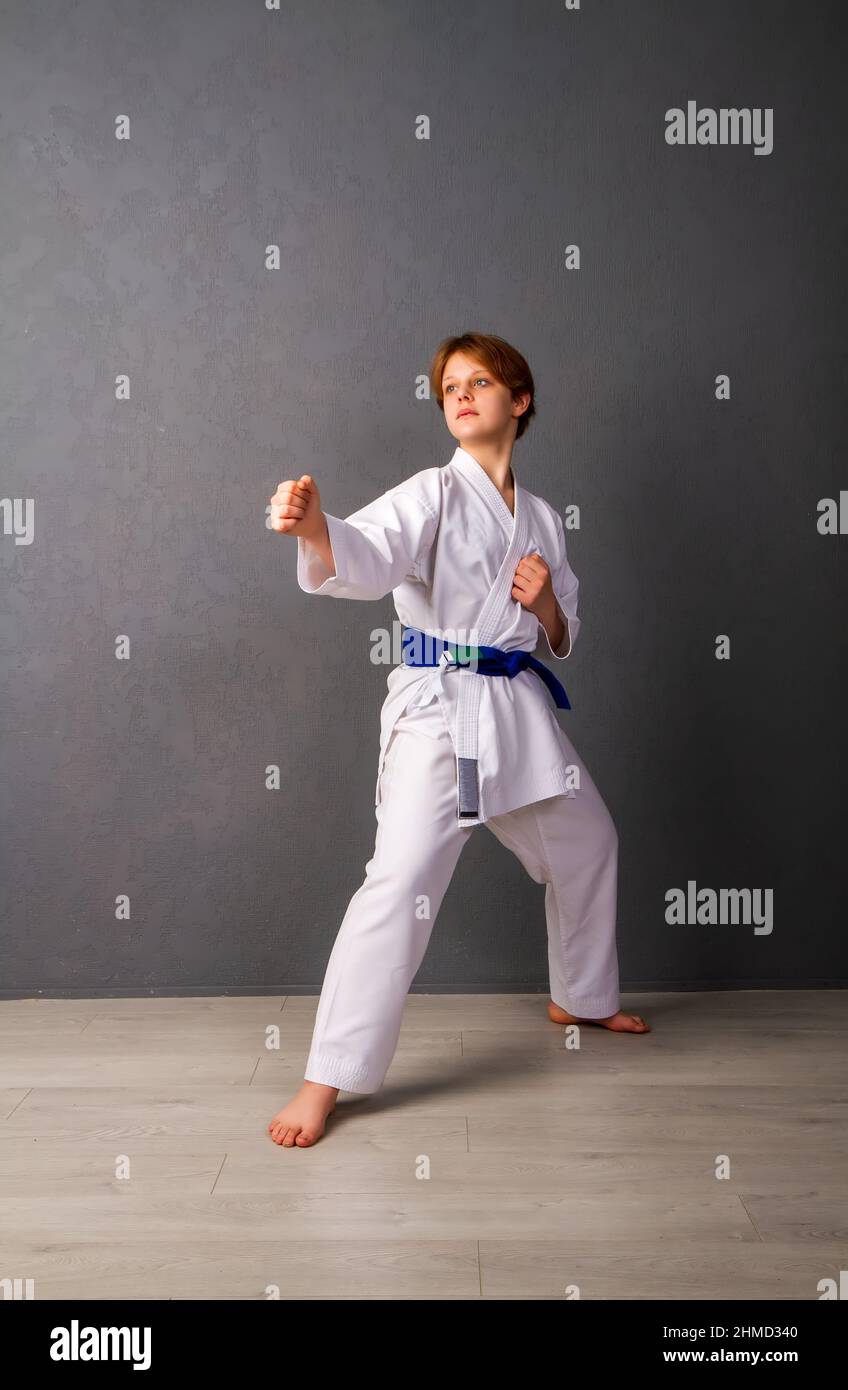 A young girl karateka in a white kimono and a blue belt trains and performs a set of exercises against a gray wall Stock Photo