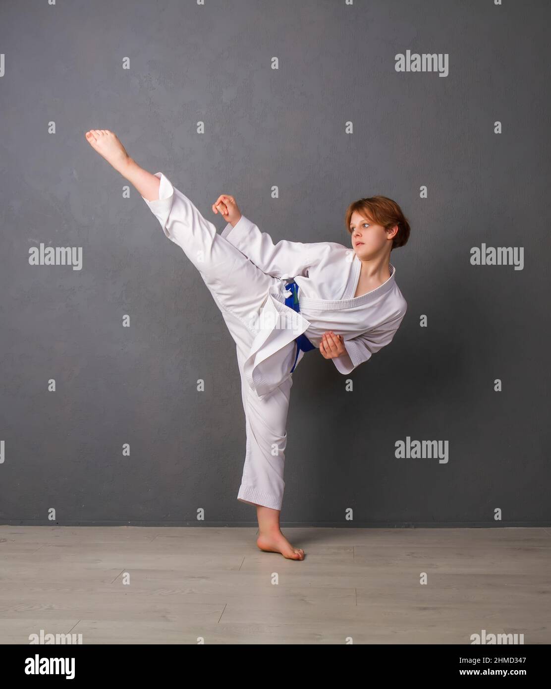 A young girl karateka in a white kimono and a blue belt trains and performs a set of exercises against a gray wall Stock Photo