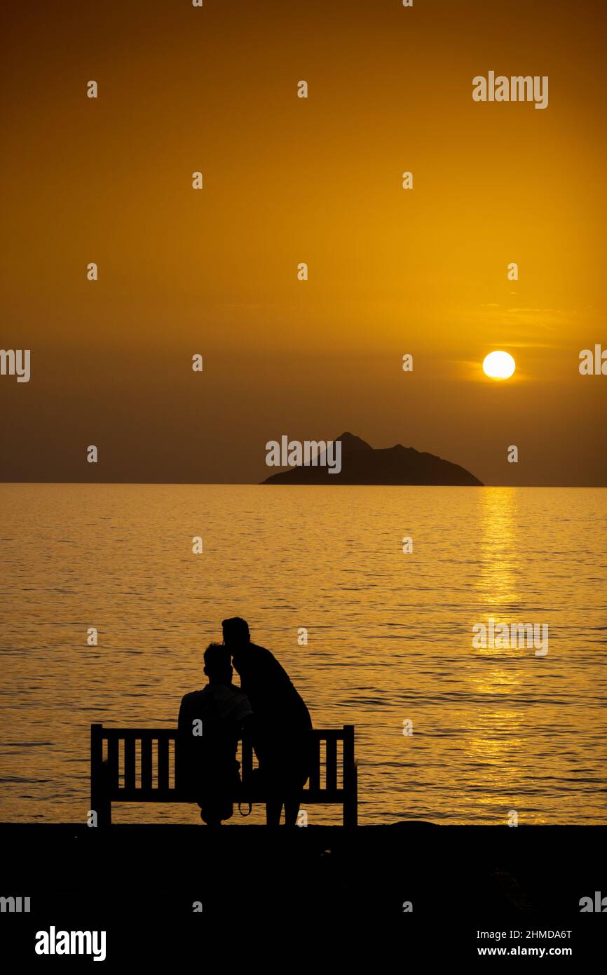 Woman kissing man sitting on bench by sea during beautiful sunset Stock Photo