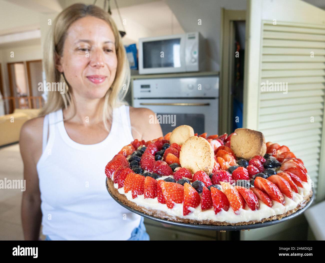 Beautiful woman holding tart garnished with whipped cream, fresh berries and cookies in kitchen Stock Photo
