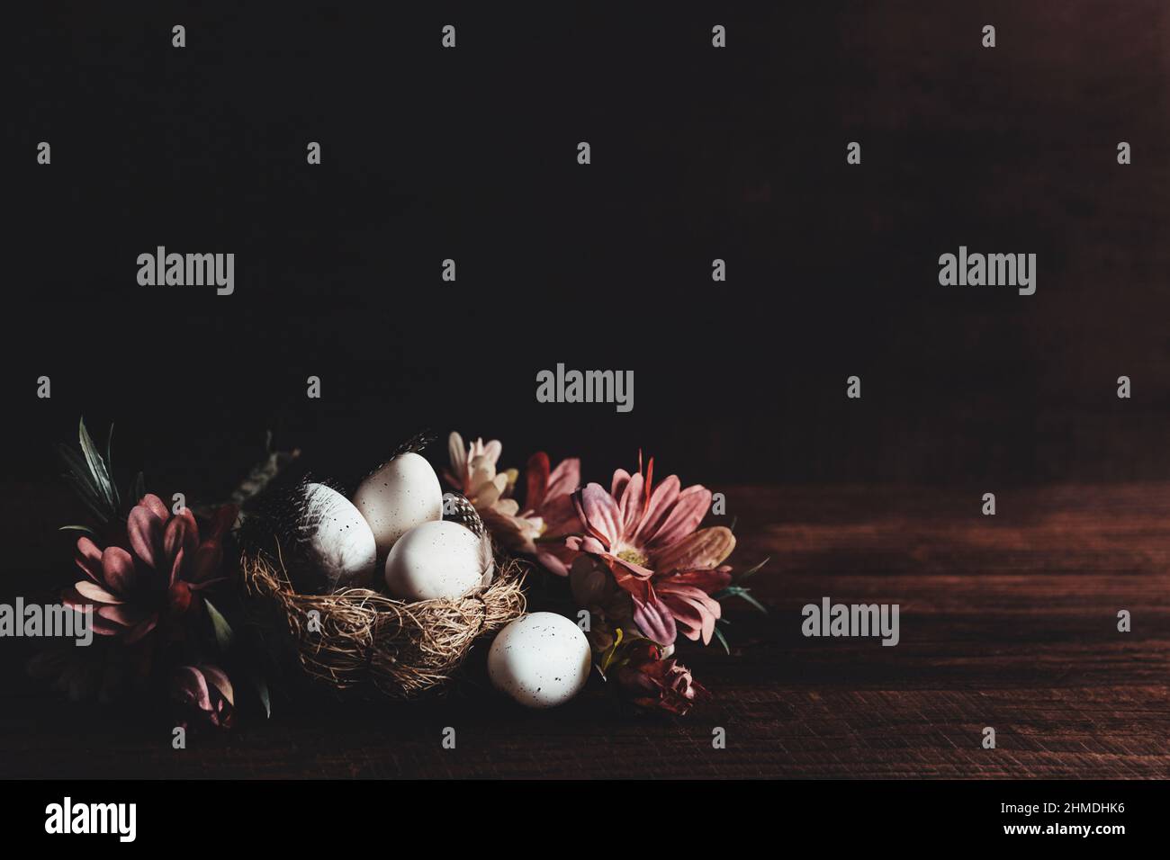 Easter holiday card with copy space to place text, mockup, wooden table background. White dotted Easter eggs in nest, pink flowers, bird feathers on wooden table. Minimal flat lay. Photo in low key Stock Photo