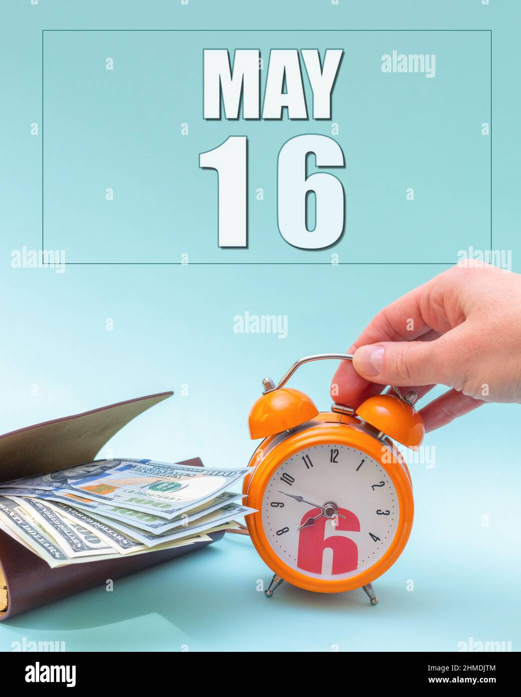 May 16th. Hand holding an orange alarm clock, a wallet with cash and a