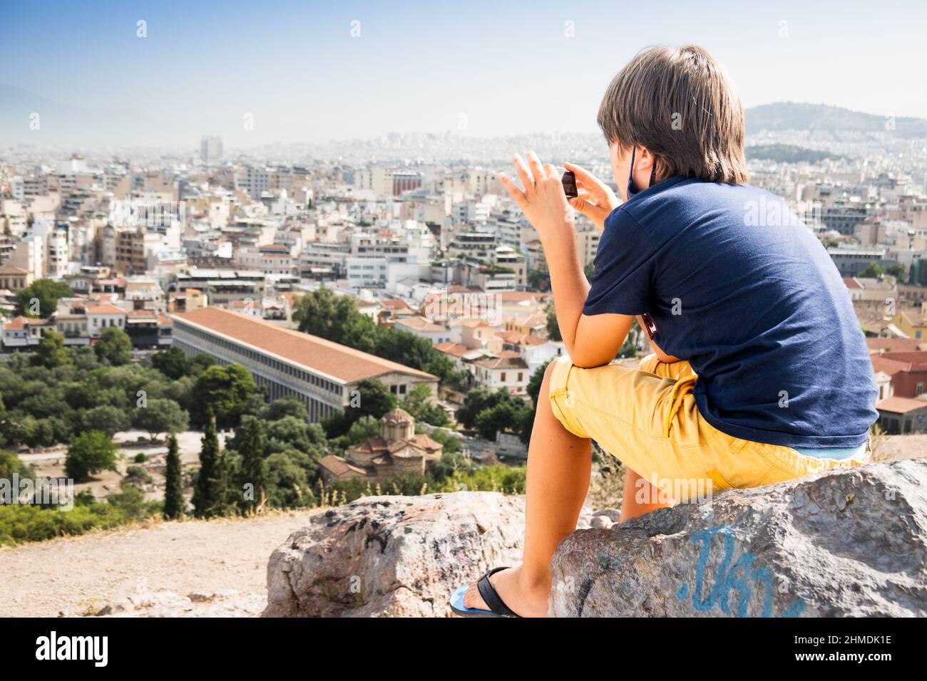 Rear view of boy sitting on stone and photographing cityscape, Athens, Greece Stock Photo