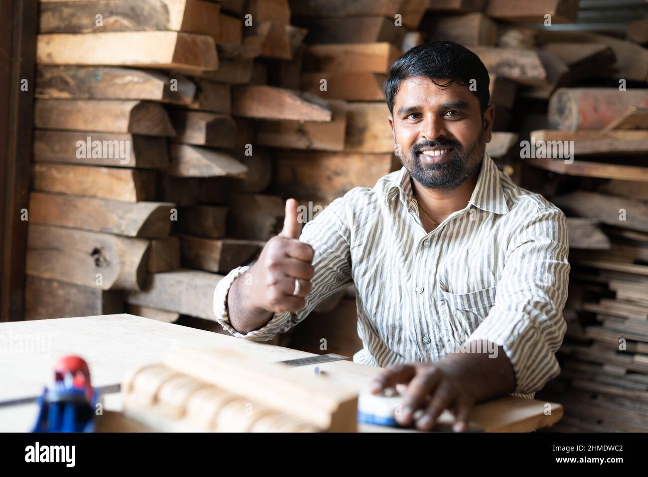 Smiling carpenter showing thumbs up by looking at camera while working at shop - concept of approve, promotion, artisan and wood working. Stock Photo