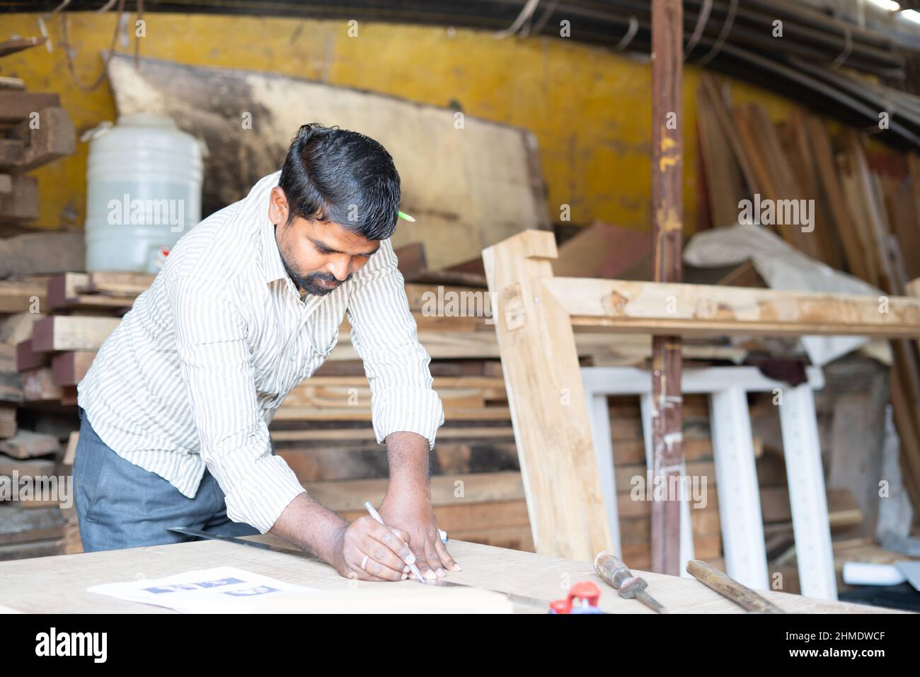 Young indian artisan marking or measuring wood by holding pencil at carpentry workshop - concept of profession woodwork, skilled occupation and hard Stock Photo