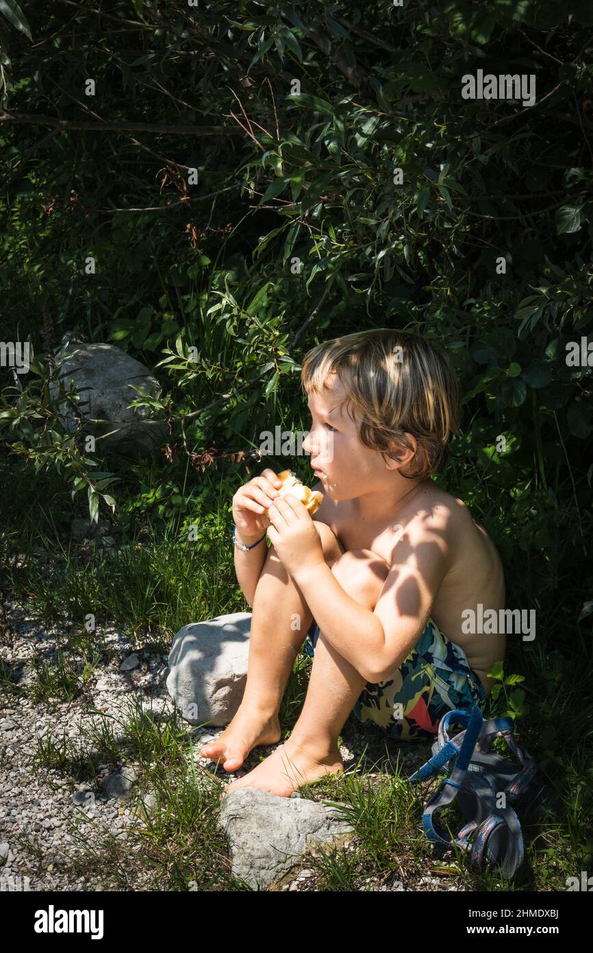 Little boy eating burger while sitting on grass under small bushes on sunny day Stock Photo