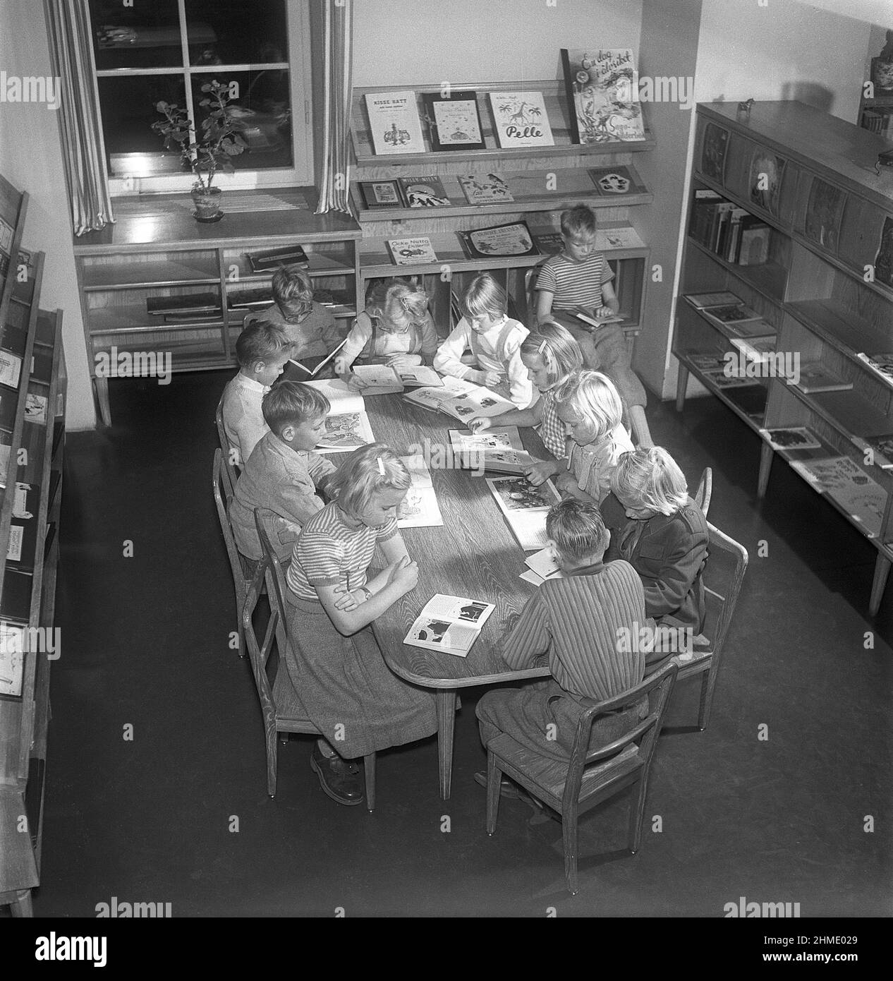 Children of the 1950s. Interior of a public library and a room with bookshelves on the walls. A group of children are sitting at a table, all reading a book each. Sweden 1951 Kristoffersson ref BE25-12 Stock Photo