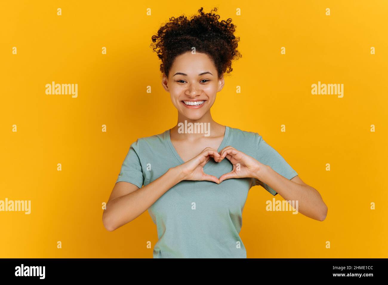 Lovely candid pleasant african american young woman, in basic t-shirt, makes heart gesture with hands, demonstrates love sign, stands on isolated orange background, looking at camera, smiling friendly Stock Photo