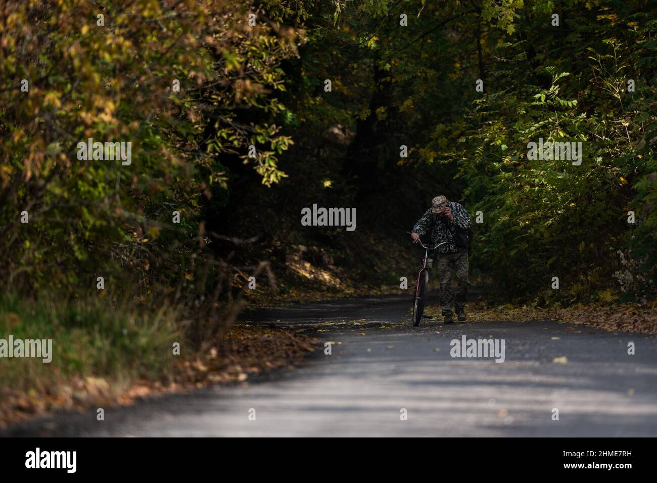 A man on the phone wearing camouflage pushes his bike up a hill in Chernobyl, near the Chernobyl Nuclear Power Plant. Stock Photo